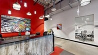 Located at 5113 S. Harper in Chicago's Hyde Park neighborhood, this single-floor commercial space offers a thoughtfully designed environment that blends industrial aesthetics with modern functionality. The reception area immediately establishes a bold visual identity, featuring a striking red accent wall adorned with colorful artwork, a weathered wood-paneled reception desk, and an array of pendant lighting suspended from exposed ceilings with visible ductwork and conduit. The patterned floor tiles in red and gray add a dynamic graphic element underfoot. Beyond the reception, an open common area provides generous communal seating with rustic reclaimed wood tables and mixed seating, ideal for collaboration or informal gatherings. Polished concrete flooring runs throughout, reinforcing the space's contemporary industrial character. A dedicated conference room features a substantial stone-topped table, ergonomic mesh chairs, a large display screen, and a whiteboard, offering a focused setting for meetings. A corridor lined with private offices or enclosed rooms extends along one side, with red accent panels and structural columns maintaining visual consistency. A comfortable lounge area with a sofa, accent chair, and patterned rug sits beside a window offering natural light and neighborhood views. A kitchenette with a clean white countertop, stainless sink, and open shelving rounds out the amenities.