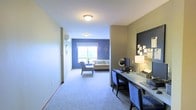 This single-floor space offers a thoughtfully arranged layout that balances comfort and functionality. A carpeted hallway leads into the main living area, where a cozy sitting room is highlighted by a bold blue accent wall and a window that draws in natural light, creating a welcoming focal point. The seating arrangement, featuring a sofa and armchair, is complemented by decorative wall accents that add a personal touch to the space. Adjacent to the entry, a dedicated workspace runs along one wall, featuring a long desk surface, ample room for a computer setup, and a large framed bulletin board — ideal for staying organized. A desk lamp provides targeted lighting for the work area. The neutral-toned walls throughout the hallway and workspace create a calm contrast to the vibrant blue of the living area, while the continuous carpeted flooring unifies the space. Flush-mounted ceiling lights provide consistent illumination throughout. The overall ambiance is practical yet comfortable, offering distinct zones for relaxation and productivity within a compact, well-utilized single-level layout.