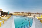 This thoughtfully designed outdoor amenity space offers a compelling blend of relaxation and social gathering areas centered around a generously sized, crystal-clear swimming pool. The pool deck is outfitted with vibrant yellow-framed lounge chairs and colorful umbrellas, providing ample space for sunbathing and leisure. A sleek metal safety fence encloses the perimeter, ensuring both security and open sightlines to the surrounding landscape and skyline.

Adjacent to the pool, a covered outdoor lounge and kitchen area extends the functionality of the space year-round. The covered zone features a modern paneled ceiling with integrated lighting and retractable elements that open toward the pool, creating a seamless indoor-outdoor connection. Multiple metal dining tables paired with teal woven chairs offer comfortable seating for groups, while a stainless steel grill and an outdoor kitchen with a sink provide everything needed for al fresco entertaining.

The overall design aesthetic is contemporary and clean, with large-format light-toned pavers underfoot and horizontal board cladding on the surrounding walls. Neatly trimmed evergreen shrubs and landscaping elements add a touch of greenery against the urban backdrop of a modern multi-story building. The space is bright, open, and airy, making it an inviting destination for outdoor dining, recreation, and relaxation.