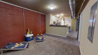 This single-floor space showcases an intriguing blend of industrial and contemporary design elements. The open-plan layout connects a spacious carpeted living area to a kitchen, creating a cohesive and functional flow throughout. A striking full-height exposed brick wall anchors one side of the main living space, lending a raw, urban character to the interior. Visible metal ductwork runs along the ceiling, reinforcing the industrial aesthetic.

The kitchen is defined by a wraparound dark-topped peninsula that serves as both a workspace and a dividing element between the living and cooking areas. Overhead, a series of teal pendant lights hang at varying heights, adding a pop of color and a modern touch to the space. The kitchen is equipped with stainless steel appliances, including a microwave and refrigerator, and features white cabinetry for ample storage.

Neutral-toned carpet covers the living area floor, softening the industrial elements and providing a comfortable underfoot feel. The overall ambiance balances utilitarian features with warm accents, resulting in a distinctive space that feels both functional and characterful.