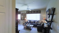 This single-floor space centers around a thoughtfully arranged living room that balances comfort and style. The room is furnished with dark leather sofas and chairs, complemented by decorative throw pillows that add texture and warmth. A ceiling fan with broad blades is mounted centrally, providing both air circulation and a subtle architectural focal point overhead.

Natural light enters through a large window dressed with vertical blinds and a decorative valance featuring a nature-themed pattern, contributing to the room's warm, earthy aesthetic. Wall art with wildlife and nature motifs reinforces this theme, creating a cohesive, rustic feel throughout the space.

A wooden coffee table sits at the center of the seating arrangement, styled with candles and decorative accents. Matching end tables flank the seating area, each topped with table lamps that provide soft, ambient lighting. A dark wood shelving unit is partially visible to one side, offering additional storage and display space.

The floor is covered in neutral-toned carpet, softening the space and tying together the warm color palette. Overall, the living room presents a comfortable and inviting atmosphere with consistent nature-inspired design details.