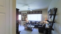 This single-floor space centers around a thoughtfully arranged living room that balances comfort and style. The room is furnished with dark leather sofas and chairs, complemented by decorative throw pillows that add texture and warmth. A ceiling fan with broad blades is mounted centrally, providing both air circulation and a subtle architectural focal point overhead.

Natural light enters through a large window dressed with vertical blinds and a decorative valance featuring a nature-themed pattern, contributing to the room's warm, earthy aesthetic. Wall art with wildlife and nature motifs reinforces this theme, creating a cohesive, rustic feel throughout the space.

A wooden coffee table sits at the center of the seating arrangement, styled with candles and decorative accents. Matching end tables flank the seating area, each topped with table lamps that provide soft, ambient lighting. A dark wood shelving unit is partially visible to one side, offering additional storage and display space.

The floor is covered in neutral-toned carpet, softening the space and tying together the warm color palette. Overall, the living room presents a comfortable and inviting atmosphere with consistent nature-inspired design details.