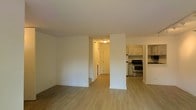 This two-floor residence offers a thoughtfully arranged layout that balances open living spaces with practical functionality. The main level features a spacious open-concept living area finished with light hardwood-style flooring that adds warmth and continuity throughout the space. Clean white walls and a neutral color palette create a bright, welcoming atmosphere.

The kitchen is partially open to the main living area and is equipped with white cabinetry, a stainless steel range, and additional appliances, offering both style and utility. A central structural column subtly defines the transition between the living and kitchen areas while maintaining the open feel of the floor plan.

A hallway connects the living space to additional rooms, providing a clear and organized flow through the home. The entry area, visible through the hallway, is well-lit with overhead lighting and includes a doorway that adds to the sense of depth and accessibility within the layout.

With its combination of open communal spaces, a functional kitchen, and a two-floor design, this residence provides a versatile and comfortable living environment suitable for a variety of lifestyles.