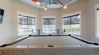 The billiard room is a single-floor amenity space designed for recreation and social gathering. At its center sits a full-size pool table with a light gray felt surface and a natural wood frame, serving as the room's focal point. A striking modern chandelier with brass arms and globe-shaped bulbs is mounted above, suspended from a decorative circular tray ceiling detail painted in bold accent colors, adding a playful, contemporary touch to the space.

Multiple large windows with roller shades line the walls, flooding the room with natural light while offering views of the surrounding landscaped grounds. High ceilings contribute to an open, airy atmosphere. Along the windowed wall, tall bistro-style tables paired with modern chairs provide seating for spectators or casual use. A dedicated cue rack holds billiard sticks and accessories, keeping the space organized and functional.

The room connects directly to an outdoor pool area, accessible via glass-paned doors with dark wood framing. The outdoor space features a generously sized swimming pool, a spa, and a sundeck furnished with lounge chairs and umbrellas. Together, the billiard room and pool area form a cohesive recreational hub suited for both active play and relaxed socializing.