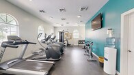 Located at 100 Piccadilly Loop in Yorktown, Virginia, this fitness center offers a dedicated exercise space designed to support a well-rounded workout routine. The room is defined by clean white walls, durable dark flooring suited for high-traffic use, and a series of elegant arched windows that line the walls, allowing ample natural light to fill the space and provide views of the surrounding outdoors.

The facility is well-equipped with a diverse selection of cardio machines, including treadmills, elliptical trainers, a recumbent bike, and a cycling bike. A multi-station cable machine serves as the centerpiece for strength training, complemented by a dedicated free weight area featuring a tiered dumbbell rack. Stability balls are neatly stored on a rack, adding flexibility training options to the mix.

A bold teal accent wall on one end of the room adds a vibrant visual contrast to the otherwise neutral interior, and a wall-mounted flat-screen television is positioned for easy viewing during workouts. A cork bulletin board is affixed near the strength equipment area, offering space for posted information and community notices. Recessed and flush-mount ceiling lighting ensures the room remains well-lit throughout. The space is accessed via a glass-paneled door and is thoughtfully organized to maximize usability across all fitness disciplines.