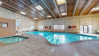 This single-floor recreational facility is designed to provide a comprehensive wellness experience, centering around a generously sized indoor swimming pool with crystal-clear blue water. The pool area features soaring ceilings with an exposed wood-paneled roof structure supported by industrial trusses, and a series of skylights and large windows that bathe the space in natural light. A hot tub sits adjacent to the main pool, and a comfortable lounge area with seating is positioned poolside for relaxation. The surrounding deck is finished in smooth concrete, offering ample space for movement around the water.

Accessible directly from the pool area through glass entry doors is a well-appointed fitness center divided into multiple zones. The cardio area is equipped with treadmills, elliptical machines, and stationary bikes, with interior windows offering views of the pool. A dedicated strength training zone houses a full rack of dumbbells, a cable weight machine, an adjustable bench, and a rowing machine. Wall-mounted flat-screen televisions are present throughout. Rubber flooring and full-length mirrors complete the functional fitness environment. The overall facility presents a clean, welcoming atmosphere suited for both aquatic and land-based exercise.