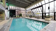 This impressive indoor pool facility is defined by its stunning glass atrium ceiling, which floods the space with abundant natural light and offers open views of the surrounding skyline and landscape. The soaring transparent roof structure, framed in dark metal, creates a dramatic architectural focal point while maintaining a warm, sun-drenched atmosphere year-round. The generously sized swimming pool features crystal-clear turquoise water and clean tile surroundings, providing an inviting environment for both leisure and lap swimming. Along the poolside deck, comfortable lounge chairs with bright yellow cushions offer relaxed seating, perfectly complemented by cheerful yellow patio umbrellas positioned over casual dining tables and chairs — ideal for unwinding or socializing. Lush potted plants and greenery are thoughtfully placed throughout the space, adding a refreshing, nature-inspired ambiance that softens the architectural lines. The exposed ceiling structure above the pool area adds an industrial yet sophisticated design element, balancing the openness of the glass atrium. Large floor-to-ceiling windows along the perimeter further enhance the sense of openness, connecting the indoor environment to the outdoor urban surroundings. Overall, this space delivers a bright, airy, and resort-like atmosphere.