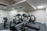 This fitness room is a dedicated exercise space designed to support a variety of cardio-focused workouts. The room is outfitted with multiple pieces of equipment, including treadmills, stationary spin bikes, an elliptical trainer, and a recumbent-style cycle, providing users with a well-rounded selection of cardio options. Patterned, multi-tone carpet flooring covers the entire space, adding a layer of comfort underfoot during workouts. A full-length wall mirror spans one side of the room, visually expanding the space and allowing users to monitor their form while exercising. The ceiling features a drop-tile panel design with integrated flat panel lighting, providing consistent, even illumination throughout. HVAC vents are positioned in the ceiling to maintain comfortable airflow. Neutral white walls keep the atmosphere clean and functional, while a small seating area with a chair offers a space to rest between sessions. The room is accessed through a single door and includes posted instructional materials on the walls. Overall, this fitness room offers a practical and functional environment for daily exercise.