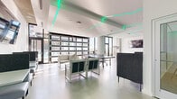 This single-floor amenity space delivers a contemporary recreational experience defined by a striking combination of modern design and versatile leisure features. The open-plan layout is anchored by polished concrete floors that extend throughout, lending the space an industrial-chic aesthetic that complements the crisp white walls and exposed structural columns.

A defining architectural feature is the geometric teal LED strip lighting embedded into the ceiling, creating a dynamic, futuristic ambiance that energizes the entire space. Large floor-to-ceiling windows and glass-panel garage-style doors flood the interior with abundant natural light, while offering views of the surrounding outdoor area.

The space is thoughtfully zoned for multiple activities. A full-size pool table in light wood tones occupies a bright corner near the expansive windows, while a regulation ping pong table provides additional gaming options. Arcade machines add a playful element along one wall. Multiple lounge seating arrangements—featuring contemporary white and charcoal upholstered furniture—offer relaxed gathering areas throughout. A wall-mounted flat-screen television enhances the entertainment offerings. The overall atmosphere is sleek, social, and purpose-built for active recreation and casual communal use.