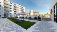 This outdoor amenity courtyard is thoughtfully designed to serve as a communal gathering space within a contemporary multi-story residential complex. The ground-level area is finished with an elegant pattern of mixed stone pavers, combining light and textured tiles to create a visually dynamic hardscape that extends throughout the space. Patches of vibrant artificial turf provide defined seating zones, furnished with modern outdoor tables, chairs, and large patio umbrellas in a fresh coastal palette of teal and neutral tones. A standout feature is the wicker pod lounge seat, offering a cozy retreat tucked against one of the perimeter garden walls. A series of sleek, charcoal-grey raised planter boxes line the boundary walls, filled with lush green shrubs that introduce natural color and greenery. A modern pergola with slatted overhead louvers provides partial shade near one of the building entrances, which is secured by a steel-framed door with wall-mounted lighting fixtures. Large floor-to-ceiling glass windows on the adjacent building facade reflect the surrounding courtyard, reinforcing the open, airy feel of the space. Stairs connect the courtyard to additional levels, and palm trees are visible beyond the perimeter walls, enhancing the tropical, resort-inspired ambiance of this well-maintained outdoor space.