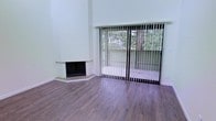 This single-floor space offers a clean, modern aesthetic defined by an open layout, generous ceiling height, and an abundance of natural light. The main living area is a standout, anchored by a stylish corner fireplace with a sleek, angular surround that serves as a focal point within the room. Rich hardwood-style flooring runs throughout, adding warmth and continuity to the space.

Large sliding glass doors fitted with vertical blinds draw the eye toward a private outdoor area, framing views of mature, verdant trees that lend a sense of tranquility and a natural connection to the outdoors. The bright white walls enhance the sense of openness, reflecting light effectively throughout the interior.

The elevated ceiling height contributes to an airy, expansive feel, making the living area feel notably spacious. The overall design balances simplicity and functionality, with clean lines and neutral finishes creating a versatile backdrop suited to a variety of décor styles. The seamless transition between the indoor living space and the outdoor area further elevates the sense of openness, making this a comfortable and inviting environment throughout the day.
