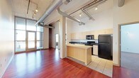 This single-floor loft-style space showcases a striking blend of industrial and modern design. The open-concept layout is defined by exposed silver ductwork running along the white ceiling, complemented by track lighting that illuminates the space efficiently. Rich, dark hardwood flooring spans the main living area, while the kitchen transitions to tile flooring, clearly delineating the two zones.

The kitchen is well-appointed with light maple cabinetry extending to the ceiling for ample storage, dark countertops, and black appliances including a refrigerator, stove, and built-in microwave. A peninsula with a breakfast bar provides additional workspace and a natural gathering point. A sink with a window view adds a functional touch.

Structural columns with brick accents add character and an authentic industrial feel. Large windows and a glass door leading to a balcony or outdoor area flood the living space with natural light, enhancing the open, airy atmosphere. The combination of raw architectural elements—exposed mechanicals, brick, and concrete—with refined finishes creates a dynamic and visually engaging living environment.