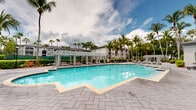 Located at 7350 NW Fourth St in Plantation, Florida, this single-story community facility offers a well-appointed outdoor retreat designed for relaxation and recreation in a lush South Florida setting. The centerpiece of the property is a generously sized resort-style swimming pool with a classic freeform shape, crystal-clear turquoise water, and a surrounding deck finished with tasteful paver stonework that provides ample space for sunbathing and leisure. A pergola structure adjacent to the pool area features a row of private cabana-style lounges with curtained enclosures, offering shaded relaxation alongside the water. Additional poolside seating, including chaise lounges and deck chairs, is thoughtfully arranged throughout the deck area. The single-story clubhouse building is finished in a clean white stucco exterior with a dark hip roof, large multi-pane windows, and open arched entryways that invite natural light and connect the interior to the outdoor environment. Manicured hedgerows, ornamental plantings, and a grove of soaring palm trees frame the entire property, creating a tropical ambiance that is both private and visually striking. Decorative paver pathways and classic lamp posts add a refined architectural touch to the grounds, enhancing the overall sense of a polished, well-maintained community amenity space.