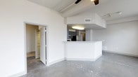 This single-floor space showcases a clean, modern aesthetic defined by polished concrete floors and a bright, open layout. The living area is anchored by exposed structural ceiling beams and a ceiling fan, lending an industrial yet refined character to the space. Abundant vertical wall space and a neutral white color palette create a sense of openness and airiness throughout.

The kitchen is seamlessly integrated into the main living area via a generous wraparound breakfast bar with a light stone-toned countertop, making it ideal for casual dining or entertaining. The kitchen itself is equipped with dark cabinetry, stainless steel appliances, and overhead lighting that keeps the workspace well-illuminated. A recessed ceiling drop above the kitchen zone subtly defines the cooking area while maintaining the open flow of the floor plan.

A doorway off the main living area leads to a walk-in closet or utility space, offering practical storage solutions. Ceiling-mounted HVAC vents and a visible ductwork rail system along the ceiling further emphasize the space's industrial design sensibility. Overall, the unit combines functional layout with contemporary finishes to create a versatile and visually engaging living environment.
