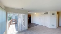 This single-story residence offers a clean, functional layout designed for comfortable everyday living. The open living area is generously sized, featuring soft wall-to-wall carpeting and large windows fitted with vertical blinds that allow abundant natural light to fill the room. A sliding glass door leads to an outdoor deck, providing a seamless connection to the exterior. The transition from the living area into the kitchen is marked by a shift to durable wood-look hard flooring. The kitchen is efficiently designed with warm-toned maple cabinetry, light countertops, a stainless steel sink beneath a window, and black appliances including a gas range and dishwasher. The bedrooms are carpeted and bright, each offering generous proportions and built-in closets with standard rod storage. One bedroom features a large window with views of surrounding greenery, while another includes a double closet for enhanced storage. The bathroom is finished in neutral tones and includes a full tub and shower combination with a curved curtain rod, white tile surround, and brushed nickel fixtures. Throughout the space, the overall aesthetic is clean and neutral, providing a versatile backdrop well-suited for personalization.