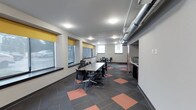 This single-floor commercial space is designed to support collaborative and functional work environments. The main area features a large rectangular meeting table surrounded by ergonomic chairs, making it well-suited for group discussions or team meetings. A secondary workstation is visible toward the rear of the space, offering additional flexibility for individual or small-group use.

The interior design embraces an industrial-modern aesthetic, highlighted by exposed metallic ductwork running along the ceiling, which adds a contemporary edge to the otherwise neutral color palette of whites and grays. Recessed and flush-mounted ceiling lights provide consistent, even illumination throughout the space.

A row of windows along one wall, fitted with yellow roller blinds, allows natural light to filter in while offering privacy and glare control. The flooring consists of a patterned carpet tile in charcoal and terracotta tones, adding visual interest and warmth to the space.

Along one wall, a kitchenette area includes a microwave and countertop surface, providing convenient amenities for occupants. The overall layout is open and well-organized, creating a professional yet approachable atmosphere suited for office or institutional use.