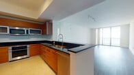This well-appointed single-floor residence showcases a contemporary design with a thoughtful layout and quality finishes throughout. The open-concept kitchen is a standout feature, boasting warm wood cabinetry, bold black granite countertops, a stainless steel appliance suite including a built-in oven, microwave, and full-size refrigerator, and a peninsula with an undermount sink that flows seamlessly into the living area. The kitchen is finished with large-format tile flooring, adding a clean, polished look. The expansive living and dining area features rich wood-tone laminate flooring, track lighting, and a bright, open feel enhanced by clean white walls and generous ceiling height. The bedrooms are carpeted for comfort and feature floor-to-ceiling sliding glass doors with vertical blinds, offering access to an outdoor balcony and views of the surrounding cityscape. The primary bathroom is elegantly appointed with a marble-topped vanity, glass-enclosed shower, soaking tub, and large-format white tile flooring. A secondary bathroom provides a clean, minimalist design with a pedestal sink and tile flooring. A washer unit is also visible within the unit, adding everyday convenience to this modern residence.