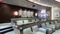 This refined, single-floor space presents a polished and professional atmosphere defined by thoughtful design and high-end finishes. The layout features tiered rows of plush cream leather chairs arranged in a theater-style configuration, each row accented with dark wood armrests and writing surfaces that lend a sense of order and comfort. The reception or service counter at the front serves as a focal point, elegantly framed by warm-toned wood paneling and a softly lit textured backdrop adorned with tasteful floral arrangements. Recessed ceiling lighting combined with subtle wall-mounted sconces creates a layered, ambient glow throughout the space, enhancing its upscale character. A mounted projector above the seating area suggests the room may serve dual purposes, functioning as both a waiting area and a presentation or briefing space. Large glass partitions on the perimeter allow natural light to filter in, maintaining an open and airy feel despite the interior setting. The overall palette of cream, dark walnut, and taupe tones contributes to a cohesive, contemporary aesthetic that conveys both professionalism and comfort, making this space well-suited for a corporate, medical, or hospitality environment.