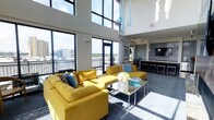 This single-floor space makes a striking impression through its dramatic double-height ceilings and expansive floor-to-ceiling windows that flood the interior with abundant natural light. The towering glass panels frame sweeping outdoor views of the surrounding cityscape and a connected balcony, reinforcing a seamless connection between the interior and exterior environments.

The open-concept living area is anchored by bold yellow sectional sofas arranged around a sleek mirrored coffee table, creating a vibrant focal point. Teal decorative accents, including cushions, glassware, and suspended teardrop-shaped pendant sculptures, complement the energetic color palette and add visual dimension to the space.

Transitioning toward the rear of the space, a kitchen and bar area features dark cabinetry, a light stone countertop, and pendant lighting overhead. Bar-height seating lines the counter, offering a casual dining or social option. The flooring throughout consists of a neutral-toned carpet that grounds the vivid furnishings.

Overall, the space balances an airy, light-filled atmosphere with contemporary design elements, making it both visually dynamic and functionally versatile.