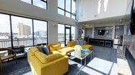 This single-floor space makes a striking impression through its dramatic double-height ceilings and expansive floor-to-ceiling windows that flood the interior with abundant natural light. The towering glass panels frame sweeping outdoor views of the surrounding cityscape and a connected balcony, reinforcing a seamless connection between the interior and exterior environments.

The open-concept living area is anchored by bold yellow sectional sofas arranged around a sleek mirrored coffee table, creating a vibrant focal point. Teal decorative accents, including cushions, glassware, and suspended teardrop-shaped pendant sculptures, complement the energetic color palette and add visual dimension to the space.

Transitioning toward the rear of the space, a kitchen and bar area features dark cabinetry, a light stone countertop, and pendant lighting overhead. Bar-height seating lines the counter, offering a casual dining or social option. The flooring throughout consists of a neutral-toned carpet that grounds the vivid furnishings.

Overall, the space balances an airy, light-filled atmosphere with contemporary design elements, making it both visually dynamic and functionally versatile.