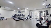 This fitness center offers a comprehensive workout environment spread across a single, open floor. The space is designed with a clean, modern aesthetic, featuring white walls, recessed lighting, and durable flooring suited for high-traffic athletic use. The main exercise area is anchored by a row of Spirit treadmills positioned in front of a large wall-mounted mirror and flat-screen television, creating an engaging cardio zone. Adjacent to this area, a cable resistance machine and free weights provide strength training options, while a dedicated elliptical room houses multiple Spirit elliptical trainers on carpet flooring for lower-impact workouts. A rowing machine and stationary cycling bike round out the cardio offerings. French doors along one wall allow natural light to filter in and provide views of the outdoors, contributing to an energizing atmosphere. A staircase with a metal railing is visible near the entrance area, alongside double glass doors that offer easy access to and from the facility. A water fountain is conveniently located near the entry. The overall layout is thoughtfully organized to accommodate a variety of fitness routines, with ample open floor space between equipment zones to ensure comfortable movement throughout the facility.