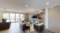 This single-floor space showcases a thoughtfully designed open living area that blends modern aesthetics with warm, inviting elements. The living room serves as the focal point, anchored by a sophisticated built-in media wall finished in a textured neutral tone. Floating wood shelves frame a wall-mounted flat-screen television, displaying curated decorative accessories that add character and depth to the space.

The seating arrangement features rich brown leather sofas and accent chairs with wooden frames and cream upholstery, centered around a layered area rug atop large-format tile flooring in a cool grey tone. An eye-catching sculptural chandelier with globe bulbs hangs overhead, complementing the recessed lighting throughout the smooth white ceiling.

Large glass sliding doors flood the interior with natural light and provide direct access to an outdoor patio area, where outdoor seating is visible beyond. The seamless connection between indoors and outdoors enhances the sense of openness. Overall, the space strikes a balance between contemporary sophistication and comfortable, livable design.