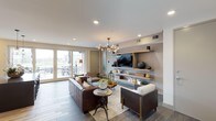 This single-floor space showcases a thoughtfully designed open living area that blends modern aesthetics with warm, inviting elements. The living room serves as the focal point, anchored by a sophisticated built-in media wall finished in a textured neutral tone. Floating wood shelves frame a wall-mounted flat-screen television, displaying curated decorative accessories that add character and depth to the space.

The seating arrangement features rich brown leather sofas and accent chairs with wooden frames and cream upholstery, centered around a layered area rug atop large-format tile flooring in a cool grey tone. An eye-catching sculptural chandelier with globe bulbs hangs overhead, complementing the recessed lighting throughout the smooth white ceiling.

Large glass sliding doors flood the interior with natural light and provide direct access to an outdoor patio area, where outdoor seating is visible beyond. The seamless connection between indoors and outdoors enhances the sense of openness. Overall, the space strikes a balance between contemporary sophistication and comfortable, livable design.