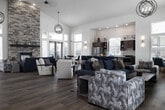 This well-appointed single-floor community space offers a generous, open-concept layout designed for both social gatherings and everyday functionality. Upon entry, visitors are greeted by a dramatic reception foyer with soaring vaulted ceilings, floor-to-ceiling windows flanking glass double doors, and an eye-catching orbital chandelier overhead. Light hardwood flooring flows continuously throughout, unifying each area seamlessly.

The main lounge is anchored by a striking floor-to-ceiling stacked stone fireplace with a dark wood mantle, serving as the architectural centerpiece of the space. Multiple seating arrangements accommodate both intimate conversations and larger group gatherings. A wall-mounted television and media console provide additional amenities near the entrance.

The fully equipped kitchen features dark cabinetry, stainless steel appliances, and a large oval island with a quartz countertop and teal upholstered bar seating. An adjacent casual dining nook, surrounded by tall windows with outdoor views, creates a bright and inviting atmosphere. A separate dining area with a marble-topped table and upholstered chairs provides an additional gathering option.

Toward the rear, a more private wing houses dedicated office or meeting rooms with glass-panel sliding doors, recessed lighting, and built-in cabinetry, offering a quiet workspace within the broader community setting.