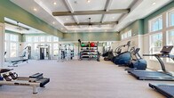 This fitness center offers a thoughtfully designed, single-level space that combines architectural elegance with comprehensive exercise functionality. The room's most distinctive architectural feature is its coffered ceiling, accented with rustic wood beams and a central ceiling fan, creating an airy, upscale atmosphere enhanced by recessed lighting throughout.

The space is organized into distinct functional zones. Along one wall, floor-to-ceiling windows flood the strength training area with natural light, offering views of an outdoor pool and surrounding landscaping. This section houses cable machines, a multi-station weight system, and free-weight benches. Full-length mirrors are strategically positioned to extend the sense of space and support proper form during workouts.

The cardio zone features a well-appointed row of elliptical trainers and treadmills, while additional equipment including rowing machines, spin bikes, and a climbing wall caters to diverse fitness needs. A dedicated rack organizes medicine balls, kettlebells, and resistance bands for functional training.

Soft sage-green walls trimmed with crisp white wainscoting contribute to the calm, motivating atmosphere. The patterned light-toned flooring throughout is both practical and visually cohesive, completing the polished, resort-style aesthetic of this well-appointed fitness facility.