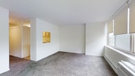 This single-floor space presents a clean, straightforward layout centered around an open living area. The room is finished with neutral grey carpeting underfoot and bright white walls throughout, creating a simple, light-filled environment. A notable architectural detail is the pass-through window connecting the living area to the kitchen, framed by warm-toned wooden cabinetry above a sink. This design element allows for visual connectivity between the two spaces while maintaining a sense of separation. A baseboard heating unit runs along one wall, providing an efficient heating solution integrated into the room's perimeter. The ceilings are smooth and white, contributing to the overall brightness of the space. The open layout of the living area offers flexibility for various furniture arrangements. The kitchen, visible through the pass-through, features honey-toned wood cabinetry and appears compact yet functional. The overall ambiance is minimal and utilitarian, with a neutral palette that provides a versatile foundation for personalization. The single-floor design ensures easy accessibility and a cohesive flow throughout the space.