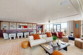 This generously proportioned single-floor suite offers a sophisticated open-plan layout that seamlessly integrates multiple functional zones within one expansive space. The interior is defined by a striking combination of terracotta hexagonal tile flooring and plush area rugs, creating a warm, layered aesthetic throughout.

The dining area anchors one end of the suite, featuring a large dark-topped table surrounded by bold geometric-patterned upholstered chairs. A statement woven chandelier hangs overhead, while the wall behind is clad in richly toned burgundy paneling with vertical stripe detailing and curated wall art, lending a refined, editorial character to the space.

Transitioning into the main lounge area, multiple seating arrangements are thoughtfully positioned to encourage both intimate conversation and broader social gatherings. Curved leather sofas, accent chairs in striped and textured fabrics, and sculptural table lamps contribute to a curated mid-century modern sensibility. A built-in media unit in deep burgundy cabinetry provides a functional focal point on one wall.

Floor-to-ceiling windows and glass-paneled double doors flood the interior with natural light while framing expansive panoramic views of the surrounding cityscape. These doors open directly onto a private outdoor terrace, extending the usable space and offering an elevated outdoor setting complete with seating and oversized planters.