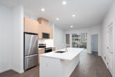 This contemporary single-floor residence showcases a clean, modern design throughout, with light wood-toned flooring and crisp white walls creating a bright, cohesive aesthetic. The open-concept kitchen serves as the heart of the space, anchored by a generous white waterfall island with an integrated sink — ideal for meal prep and casual dining. The kitchen is outfitted with warm wood-finish cabinetry, a full suite of stainless steel appliances including a refrigerator, electric range, and built-in microwave, as well as a white tile backsplash that adds a polished finishing touch. Recessed lighting illuminates the space evenly, enhancing the airy feel. The kitchen flows seamlessly into the living area, which benefits from a large sliding glass door that brings in natural light and offers a view of lush greenery outside. The bedroom is a tranquil retreat featuring a wide picture window overlooking a landscaped outdoor area, with a ceiling fan providing year-round comfort. The full bathroom is thoughtfully designed with a dual-sink vanity topped in white, warm wood cabinetry with dark hardware, a large mirror, and a soaking tub with shower. Louvered closet doors and ample storage round out this well-planned, move-in-ready space.