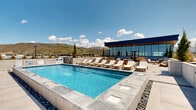 This expansive rooftop amenity deck offers a well-appointed outdoor retreat designed for relaxation and socializing. At its centerpiece is a large, rectangular swimming pool with crystal-clear water, framed by a clean concrete surround and decorative river rock borders that add a refined natural touch. Rows of teak-framed lounge chairs with plush white cushions line both sides of the pool, providing ample space for sunbathing throughout the day.

Adjacent to the pool, a spacious lounge and seating terrace features teak armchairs, club chairs, and bistro-height tables arranged to encourage gathering and conversation. A glass-walled indoor amenity building anchors one end of the deck, providing a sheltered complement to the open-air space with a bold navy fascia that adds a contemporary architectural accent.

The rooftop setting delivers breathtaking panoramic views in every direction — from rugged, arid hillsides to sweeping vistas of snow-capped mountain ranges stretching across the horizon. Slender evergreen trees are thoughtfully placed throughout the deck, adding vertical greenery and a sense of privacy. The broad open-sky exposure ensures abundant natural light throughout the day, creating a bright, resort-like atmosphere ideal for outdoor living.