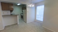 This single-story space features an open layout that connects the dining area and kitchen seamlessly. The dining area is anchored by a decorative multi-bulb chandelier and benefits from natural light streaming through a large window fitted with horizontal blinds. Neutral-toned carpet covers the dining area floor, contributing to a warm, understated ambiance. A half-wall peninsula with a countertop overhang serves as the transition point between the dining space and the kitchen. The kitchen features tile flooring, dark wood cabinetry, a freestanding gas range, and a full-size refrigerator. Recessed fluorescent lighting illuminates the kitchen area. The overall color palette is light and neutral throughout, with white walls and white trim providing a clean, straightforward aesthetic. The open sightlines between the kitchen and dining area create a practical, connected flow suited for everyday living and casual dining.