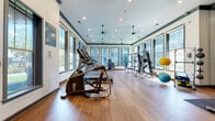 This fitness center offers a generously sized, single-floor layout designed to accommodate a comprehensive range of workout activities. The space is defined by its warm hardwood-style flooring, which extends throughout and provides a clean, durable surface suited for both equipment use and open-floor exercises.

Large windows line multiple walls, allowing abundant natural light to fill the interior while offering views of the surrounding outdoor greenery. This combination of light and sightlines contributes to an open, energizing atmosphere. Recessed lighting and pendant ceiling fixtures supplement natural light, ensuring consistent illumination throughout the space.

The fitness center is equipped with a variety of cardio machines, including treadmills, ellipticals, a stair climber, and a rowing machine, arranged to allow ample circulation. The strength training area features cable machines, a multi-station weight system, a bench press setup, and an assortment of medicine balls and free weights. A full-length mirror spans one wall, enhancing the sense of depth and supporting form monitoring during workouts.

Decorative details, including dark green crown molding and a textured accent wall, add visual character. Ceiling fans provide additional air circulation, while informational boards are posted near the entrance. The overall design balances functionality with a polished, well-maintained aesthetic.