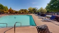 This outdoor amenity area at 2960 E North St in Greenville, South Carolina, offers a well-appointed pool and patio space designed for relaxation and leisure. The centerpiece is a generously sized swimming pool with clear turquoise water, accessible via stainless steel entry rails and marked with depth indicators for safety. The surrounding concrete pool deck is furnished with multiple woven strap lounge chairs and seating arrangements, providing ample space for sunbathing and socializing under the open sky.

Adjacent to the pool, a raised deck and patio area feature wrought iron tables paired with woven chairs, shaded by large teal umbrellas. This seating zone is enclosed by a painted wood railing with white balusters, complemented by raised wood planter borders that add a touch of greenery and visual structure to the space. Lush mature trees and well-maintained landscaping with ornamental shrubs line the perimeter, providing natural privacy and shade.

The space is enclosed by a black iron fence, with a nearby building featuring traditional siding and multi-pane windows visible in the background. The overall ambiance is bright, open, and inviting, making it well-suited for warm-weather outdoor enjoyment.