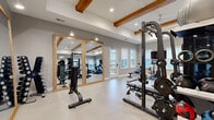 Located at 9000 Palm Passage Loop in Summerville, SC, this fitness center offers a thoughtfully designed, multi-zone workout facility spread across a single generous floor. The space is divided into three distinct areas, each tailored to a different style of exercise.

The main strength training area is the architectural centerpiece, featuring striking exposed wooden ceiling beams, recessed lighting, and ceiling fans that lend a warm yet contemporary feel. Large windows along the walls invite natural light and provide views of the surrounding community. The room is furnished with a comprehensive selection of strength equipment, including a multi-station cable machine, Smith machine, squat rack, weight benches, free weights, and battle ropes, all reflected in large wood-framed mirrors that enhance the sense of space.

A dedicated cardio zone accommodates treadmills, stationary bikes — including a Peloton — and a rowing machine, alongside a wall-mounted flat-screen TV, making it ideal for endurance-focused workouts.

The studio room offers a clean, bright environment with full-length wall mirrors, a ballet barre, soft flooring, and a mounted TV, creating an inviting space for cycling, stretching, or group-style workouts. An adjacent room with spin bikes and a ceiling fan rounds out the facility, providing a quieter setting for focused cardio sessions. Together, these zones create a comprehensive and welcoming fitness destination.