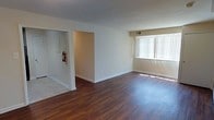 This single-floor unit features a spacious, open-concept living area finished with rich, dark hardwood flooring that extends throughout the main space. The room is bathed in natural light streaming through a large window fitted with vertical blinds, creating a bright and welcoming atmosphere. Neutral-toned walls in soft beige and white complement the warmth of the flooring, contributing to a clean, understated aesthetic. Dual HVAC vents are positioned along the upper wall and ceiling, ensuring consistent climate control throughout the space. A hallway off the main living area provides access to additional rooms, with a fire safety panel and extinguisher mounted near the entry — a practical detail indicating attention to safety standards. The layout offers a generous sense of openness, with clear sightlines across the room that lend themselves well to flexible furniture arrangements. White baseboards frame the perimeter, adding a crisp finishing touch to the overall interior. The combination of natural light, hardwood floors, and a straightforward open layout makes this a functional and comfortable living space.