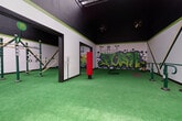 This single-level fitness facility is designed with an energetic, urban aesthetic that sets it apart from conventional gym spaces. The flooring throughout is covered in bright green artificial turf, creating a cohesive, sporty atmosphere while providing a practical surface for diverse workout activities. The space is divided into interconnected zones accessible through wide, black-framed open doorways, allowing for seamless movement between areas.

The main training area features an array of green and gold outdoor-style exercise stations, including pull-up bars, suspension rigs, gymnastic rings, and resistance equipment. A red freestanding punching bag and thick battle ropes are also present, catering to combat and conditioning training. The walls are finished in white with bold green accent stripes, and one wall showcases an eye-catching graffiti mural depicting a muscular figure above the word "FITNESS," while another features a vibrant "PUSH" graffiti piece surrounded by graphic elements.

The ceiling transitions from a dark painted upper section to a translucent corrugated roof panel that allows natural light to filter in, while industrial wall-mounted fans ensure airflow. A separate enclosed area with a gated exterior access point and chain-link fencing above connects the space to the outdoors, with surrounding trees visible beyond the perimeter.