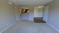 This single-floor residence is designed with an open-concept layout that promotes a seamless flow between living spaces. The main living area is finished with light-toned wall-to-wall carpeting, creating a comfortable and cohesive feel underfoot. Neutral gray-toned walls run throughout, providing a versatile backdrop suitable for a variety of interior styles.

The kitchen is positioned centrally and accessible via a raised pass-through counter, which serves as both a functional workspace and a visual divider between the kitchen and the main living area. The kitchen is equipped with stainless steel appliances, including a refrigerator and stove, and features dark cabinetry with a pendant light overhead that adds a focal point to the space. The pass-through design keeps the kitchen connected to the living area without fully opening the two spaces.

Adjacent to the main living area is a secondary room distinguished by its dark hardwood-style flooring, offering a natural contrast to the carpeted zones. This room is accessible through a white paneled door and benefits from ceiling-mounted lighting. White baseboards throughout the unit provide clean, finished transitions between the walls and flooring.