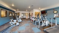 Located at 4924 Shelby Ave in Rapid City, South Dakota, this fitness center offers a comprehensive exercise environment within a single, open-plan room. The space is thoughtfully laid out to accommodate a wide variety of workout routines, making efficient use of its footprint.

The room features a two-tone modular carpet floor in grey and blue, providing a clean and functional surface throughout. Recessed lighting in the white ceiling keeps the space bright and well-lit, while multiple windows along the walls allow natural light to fill the room and offer views of the surrounding area.

Cardio equipment is prominently featured, including several treadmills, elliptical trainers, and upright and recumbent exercise bikes, all arranged to allow comfortable spacing between users. A dedicated free weights area includes a full dumbbell rack alongside a weight bench and exercise balls, catering to strength training needs. A multi-station cable machine provides additional resistance training options.

A large wall mirror along one side visually expands the space and supports proper form during workouts. The walls are accented with a teal-blue trim detail, adding a pop of color to the otherwise neutral palette. A motivational wall decal completes the atmosphere. Entry to the fitness center is through a set of interior doors, and an AED and safety signage are visible near the entrance.