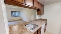 This single-floor residence offers a practical and well-appointed layout designed for comfortable everyday living. The kitchen serves as a functional centerpiece, featuring rich warm-toned wood cabinetry that extends both above and below the countertops, providing generous storage throughout. The countertops display a granite-style surface in earthy tones, complemented by a stainless steel sink with a classic faucet and a dishwasher seamlessly integrated into the cabinetry. The kitchen opens directly into the living area, creating a connected, flowing space ideal for both daily use and casual entertaining. The living area is carpeted and benefits from natural light streaming through a window that frames a view of lush greenery outside, lending a warm and inviting atmosphere to the space. The flooring transitions from carpet in the living area to tile or hard flooring in the kitchen, offering a clean distinction between the two zones. A bathroom is also present within the unit, as visible in the overview, maintaining the home's clean and neutral aesthetic. The overall color palette of light walls and warm wood tones creates a cohesive, bright, and welcoming environment throughout the single-level floor plan.
