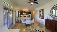 This single-floor residence offers a seamless open-concept layout that integrates the living, dining, and kitchen areas into one cohesive and inviting space. The polished concrete floors reflect light throughout, lending the interior a sleek, modern aesthetic that ties the entire floor plan together.

The kitchen is well-appointed with dark espresso cabinetry, stainless steel appliances including a French door refrigerator and range, a tile backsplash, and granite or stone countertops. A raised breakfast bar provides casual seating with bar stools, making it a practical and social hub of the home. Pendant lighting hangs above the kitchen area, adding warmth and a designer touch.

The dining area sits adjacent to the kitchen and features a round dining table surrounded by elegant white upholstered chairs, creating a refined yet comfortable setting for meals and entertaining. A ceiling fan with a light fixture is centrally positioned overhead, contributing both function and style to the space.

Wall art and carefully chosen accessories, including a zebra-print area rug in the living zone, add personality and visual interest. The layout allows for natural light to filter through, enhancing the bright and airy feel of the interior. Overall, the space presents a polished and cohesive living environment with contemporary design sensibilities throughout.