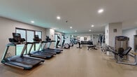 Located on Euclid Avenue in the heart of Cleveland, Ohio, this fitness center spans a generous single floor and is divided into two distinct exercise rooms, offering a comprehensive wellness environment. The main workout area features durable speckled flooring and is outfitted with an extensive selection of equipment, including treadmills, ellipticals, spinning bikes, cable machines, selectorized strength stations, free weights, and adjustable benches — providing options for both cardio and resistance training. Recessed lighting illuminates the space evenly, while large mirrors along the walls enhance the sense of openness. A dedicated studio room offers a clean, versatile setting with light hardwood-style flooring, full-length mirrors, and exposed white brick accent walls that add a touch of industrial character. This studio is stocked with exercise mats, stability balls, resistance accessories, and wall-mounted storage racks for medicine balls and fitness tools, making it well-suited for yoga, stretching, and group exercise. A transitional area with built-in shelving and glass partitions connects the rooms, providing convenient storage and a clear sightline throughout the facility. The overall design is modern and functional, maintaining a bright, clean aesthetic throughout.
