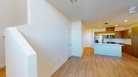 This two-story residence is designed with a clean, contemporary aesthetic throughout. The main level features light wood-tone flooring that runs through an open-concept layout connecting the entryway, living area, and kitchen. The kitchen is a standout feature, equipped with warm wood-grain cabinetry, stainless steel appliances including a French door refrigerator and range, and a large central island finished with a marble-look quartz countertop. A sleek linear pendant light hangs above the island's undermount sink, complementing the modern design. A convenient half bath on the main level includes a pedestal sink and wood-tone flooring. A dedicated laundry room houses side-by-side washer and dryer units with built-in shelving. A bright, clean hallway with recessed lighting connects the main living spaces. The upper level transitions to plush carpet flooring and features a generously sized bedroom filled with natural light from multiple windows, a ceiling fan, and access to a walk-in closet with dual hanging rods. Throughout, the space maintains a neutral, light-filled palette that enhances its open and airy ambiance.