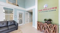 The Willows at Little Egg presents a thoughtfully designed single-story space that immediately conveys a relaxed coastal aesthetic upon entry. The lobby serves as the centerpiece of the property, featuring dramatically high ceilings that amplify the sense of openness and airiness throughout the space. Soft aqua and sage green walls establish a soothing, nature-inspired color palette that carries throughout the interior.

A standout architectural detail is the prominent feature wall rendered in fresh green, boldly displaying The Willows branding in vibrant pink lettering — creating a striking focal point for visitors. Beneath it sits a distinctive console table crafted from intertwined driftwood branches topped with a glass surface, reinforcing the organic, coastal character of the space.

White-trimmed doors with louvered blinds lead to additional rooms, offering a clean contrast against the soft wall tones. Light-toned flooring contributes to the bright, welcoming ambiance. A comfortable seating area with a charcoal chair provides a relaxed waiting space, while recessed lighting and elevated transom windows allow natural light to filter in, enhancing the overall airy and inviting atmosphere.