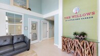 The Willows at Little Egg presents a thoughtfully designed single-story space that immediately conveys a relaxed coastal aesthetic upon entry. The lobby serves as the centerpiece of the property, featuring dramatically high ceilings that amplify the sense of openness and airiness throughout the space. Soft aqua and sage green walls establish a soothing, nature-inspired color palette that carries throughout the interior.

A standout architectural detail is the prominent feature wall rendered in fresh green, boldly displaying The Willows branding in vibrant pink lettering — creating a striking focal point for visitors. Beneath it sits a distinctive console table crafted from intertwined driftwood branches topped with a glass surface, reinforcing the organic, coastal character of the space.

White-trimmed doors with louvered blinds lead to additional rooms, offering a clean contrast against the soft wall tones. Light-toned flooring contributes to the bright, welcoming ambiance. A comfortable seating area with a charcoal chair provides a relaxed waiting space, while recessed lighting and elevated transom windows allow natural light to filter in, enhancing the overall airy and inviting atmosphere.