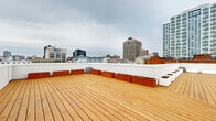Located in the heart of downtown New Haven, Connecticut, this rooftop deck at 29 Crown Street offers a generously sized open-air retreat with striking panoramic views of the city skyline. The deck's layout follows an L-shaped configuration, maximizing usable outdoor space and providing multiple vantage points across the urban landscape, including prominent downtown high-rises and the broader cityscape extending to the horizon.

The flooring consists of warm-toned, natural wood planking laid in clean linear rows, lending the space an inviting and polished aesthetic. Surrounding the perimeter are a series of rectangular planter boxes in both crisp white and rich wood finishes, offering the potential for greenery and adding a refined, landscaped character to the rooftop environment. Low white corrugated parapet walls define the boundary of the deck, providing safety while maintaining unobstructed sightlines.

A central rooftop mechanical enclosure with access doors is integrated into the design, finished in matching white paneling that keeps the overall look cohesive. Wall-mounted lighting fixtures are positioned along the structure, supporting use of the space beyond daylight hours. The open-sky setting ensures abundant natural light throughout the day, making this rooftop deck a distinctive and functional outdoor space in the midst of New Haven's vibrant urban core.