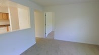 This single-floor space presents a clean, minimalist layout finished in neutral white tones throughout. The main living area features wall-to-wall beige carpeting, providing a warm and understated foundation for the room. Smooth white walls and white baseboards contribute to a bright, airy atmosphere, while the overall design maintains a simple, functional aesthetic.

A notable architectural feature is the rectangular pass-through window cutout built into the left wall, which connects this room to an adjacent area — likely a kitchen or utility space — allowing light and communication to flow between the two zones. The transition between the carpeted living area and the adjoining space, visible through the open doorway, shows a change to hard flooring, suggesting a distinct functional separation between rooms.

The open doorway leads into a connecting hallway or room, adding a sense of depth to the layout. White-painted door trim and clean lines throughout reinforce the space's straightforward, practical character. Electrical outlets are positioned along the walls for functional convenience. Overall, the space offers a neutral, move-in-ready canvas well-suited for a variety of uses and furnishing styles.