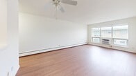 This 1-bedroom, 1-bathroom unit in Monona, Wisconsin offers a practical and comfortable layout across a single floor. The living room is bright and open, featuring warm hardwood-style flooring, a ceiling fan, baseboard heating, and a large window fitted with an in-unit AC unit that fills the space with natural light. The open layout connects seamlessly to the kitchen, which features warm wood-toned cabinetry, a white appliance suite, and a pass-through counter that enhances the sense of connectivity between rooms. The bedroom is a comfortable size, finished with plush neutral carpeting, a ceiling fan, and a row of horizontal windows that allow in soft, diffused light. A spacious sliding-door closet provides ample storage. The bathroom is a standout feature, showcasing classic retro character with sage green tile wainscoting, a mosaic tile floor, a white vanity with wood cabinet base, and a full bathtub with shower combination. Throughout the unit, crisp white walls and clean lines create a bright, neutral backdrop that is easy to personalize. Baseboard heating is present throughout, ensuring comfort during Wisconsin winters. The overall ambiance is clean, functional, and move-in ready.